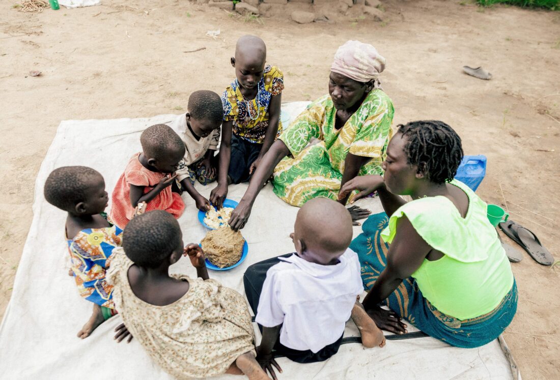 Vrouw en kinderen zitten op de grond en maken samen kleifiguren in een open ruimte in Afrika.