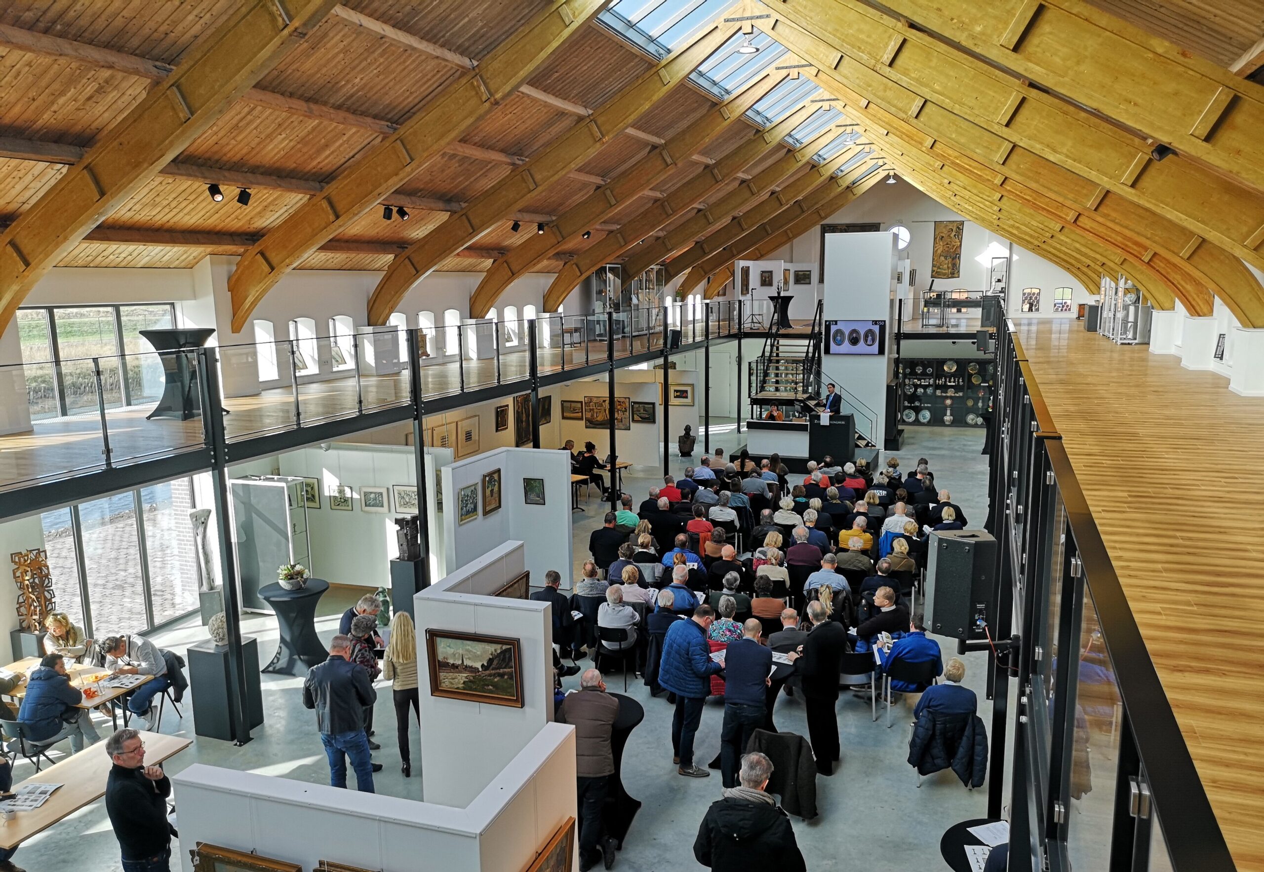 Evenement in een moderne zaal met houten plafond en grote ramen met publiek en presentatietafel.