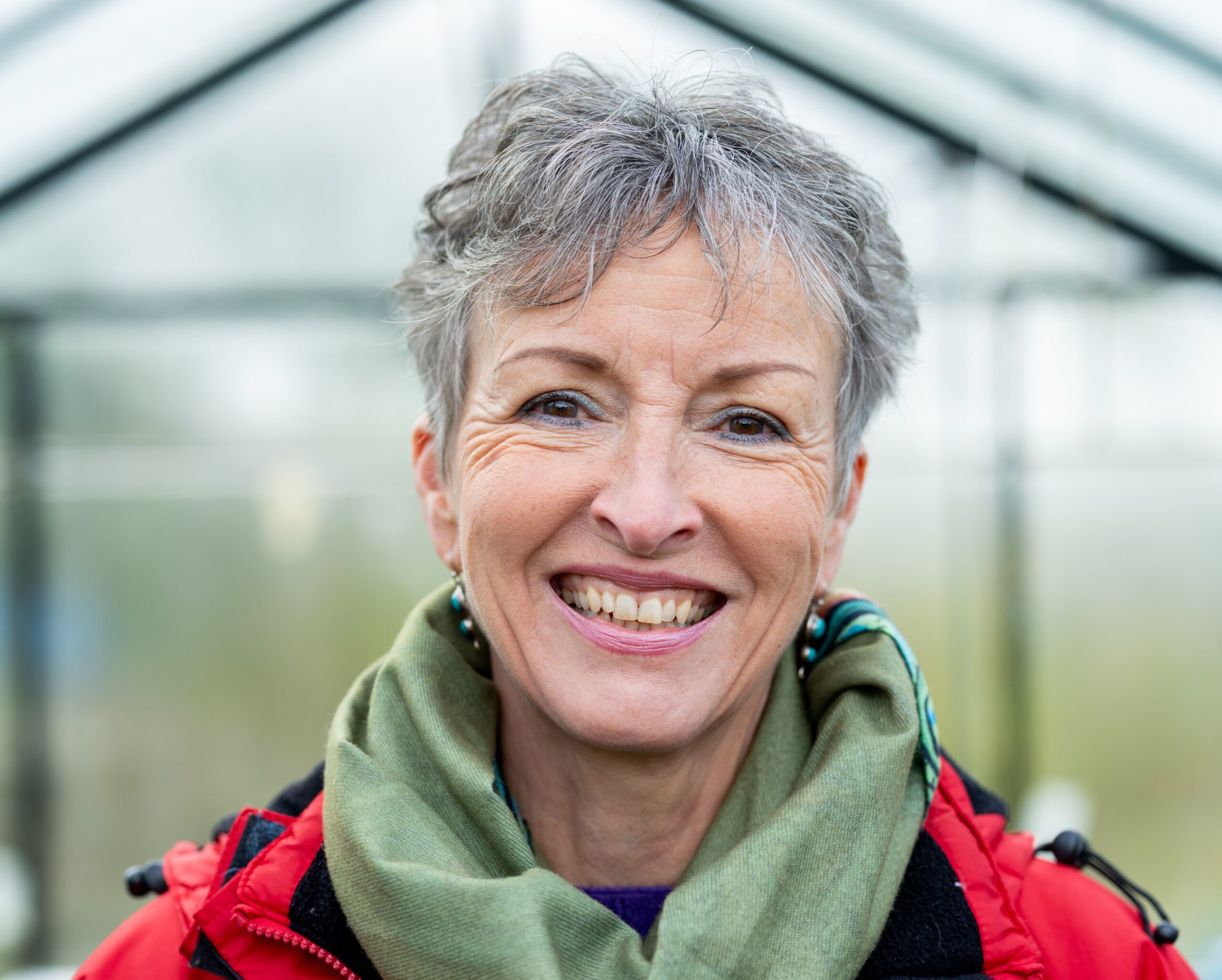 Vrolijke vrouw met kort grijs haar in een kas of serre met groene achtergrond.