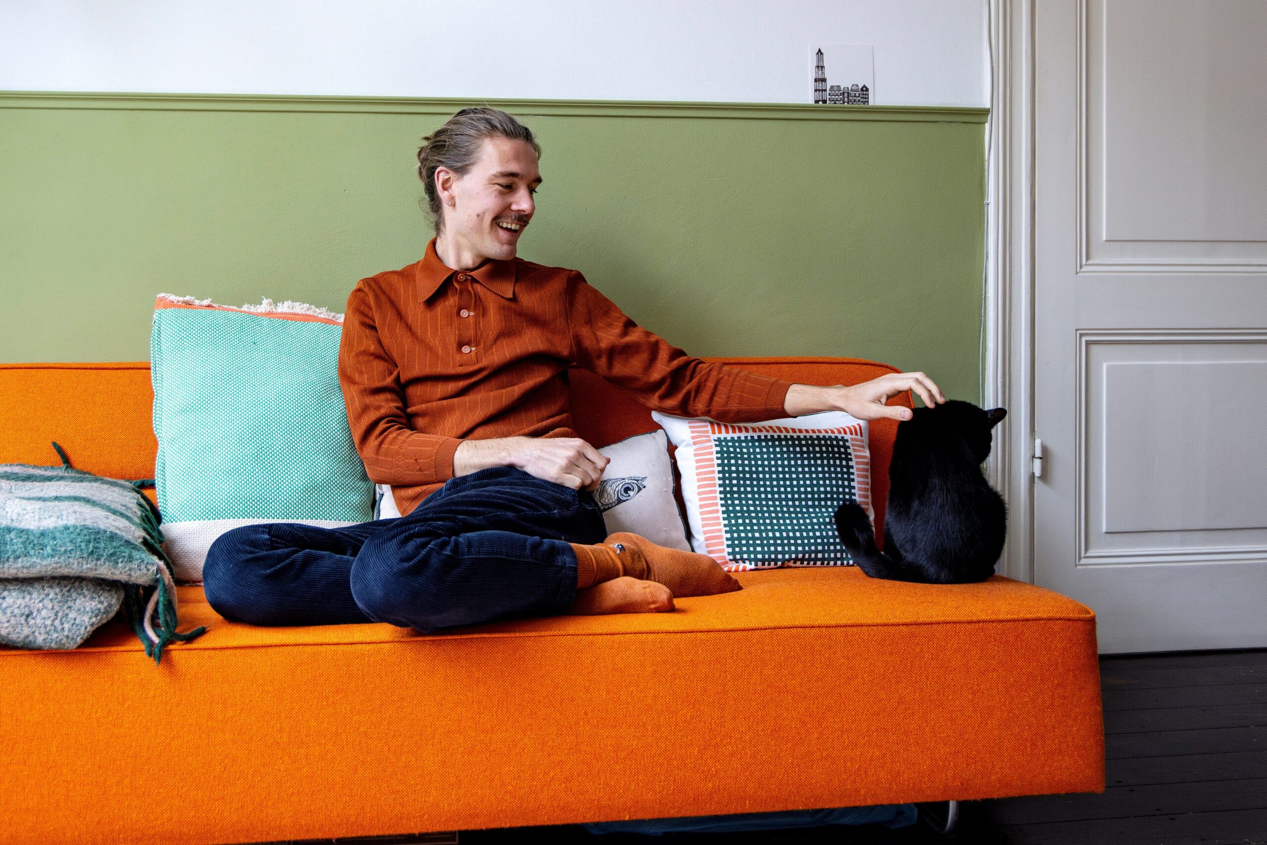 Vrouw aait kat op oranje bank in woonkamer met groene muur en decoratieve kussens.