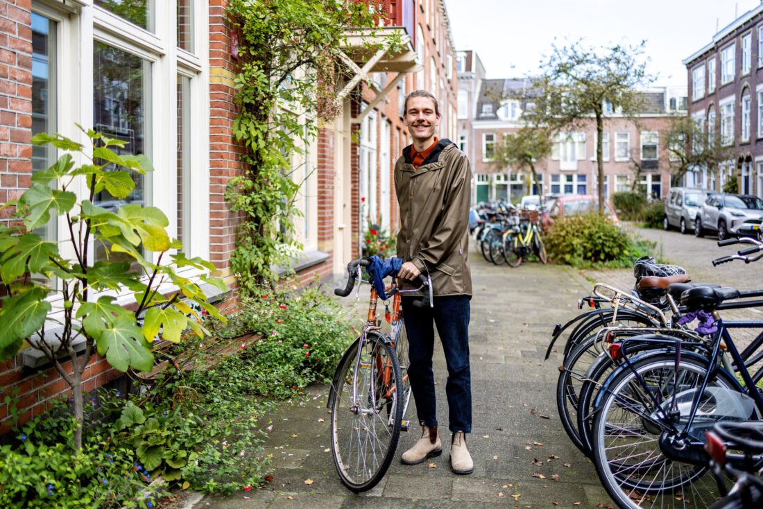 Man met fiets op straat in een woonwijk met fietsenrekken en groene planten naast een gebouw.