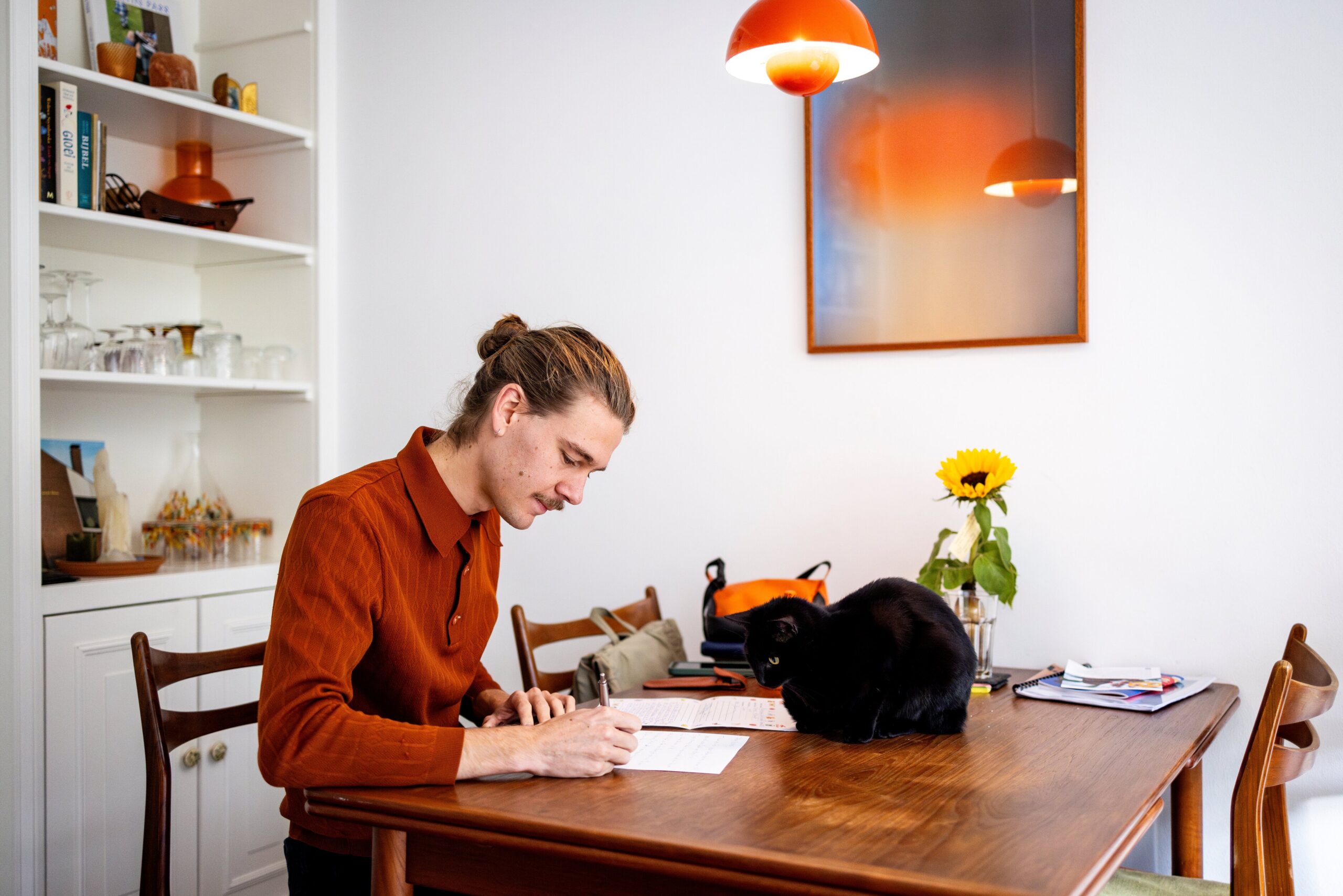 Vrouw schrijft op papier aan tafel met kat en bloemen in de achtergrond in een lichte keuken.